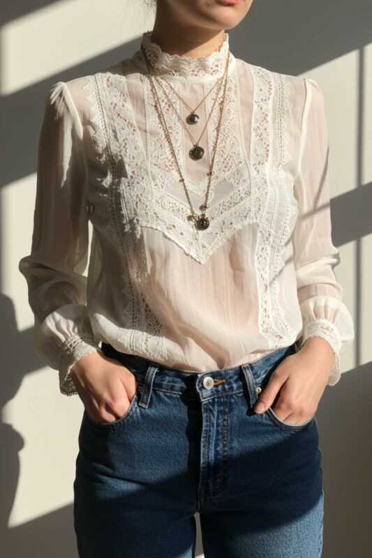 White vintage-inspired sheer blouse with lace details styled with dark denim and layered jewelry.
