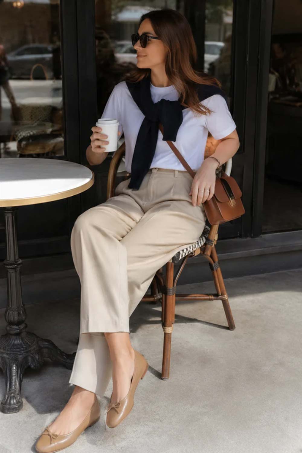 Woman in white cotton t-shirt, beige pleated trousers, navy cashmere sweater draped over shoulders, ballet flats demonstrating third-piece styling technique