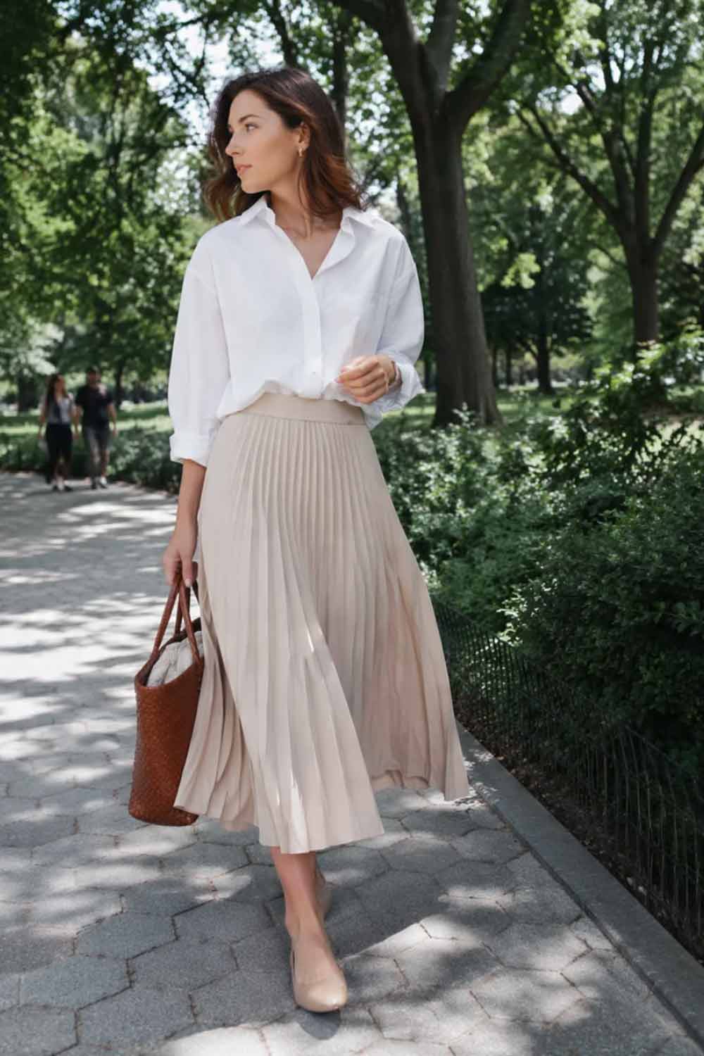 Woman wearing an oversized white poplin button-down shirt tucked into a beige pleated midi skirt with tan leather ballet flats and a woven tote bag